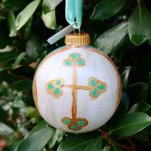 Load image into Gallery viewer, An original hand painted cross on a white ornament. The gold cross has dots of green and blue on the ends. The abstract design features blues, white, green and gold with a gold top and a green ribbon.  It is displayed hanging on a tree.
