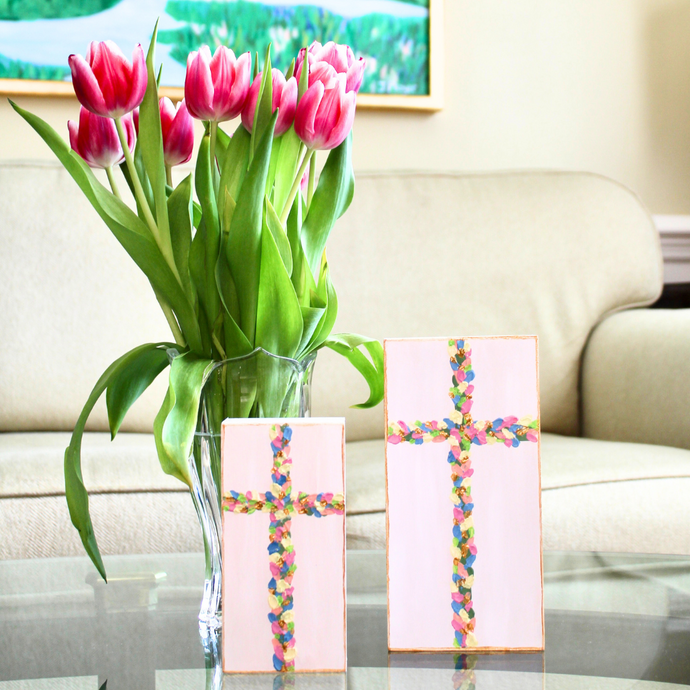 Flowered Crosses are original works of art on wood panels. These two crosses are vertical artworks in two different sizes. They have shades of pink, yellow, blue, coral, gold and green on a white backrground. The edge is outlined in gold. They sit on a table with a vase of flowers beside them and a sofa behind them.