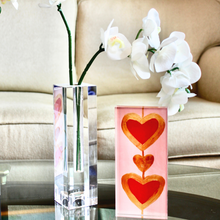Load image into Gallery viewer, Heart Art. A 2. 5 x 7.5 vertical free standing acrylic block with a pink background and two red hearts outlined in gold connected by a gold line. It istyled on a mirrored tray beside a glass vase holding orchids.