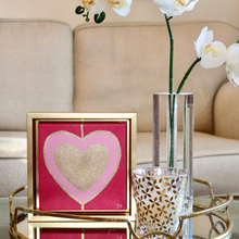 Load image into Gallery viewer, Heart Art Acrylic on Canvas. Gold and pink hearts on red background. This square heart pop art painting comes in a gold frame and is dislplayed by a vase of orichids with a sofa in the background. Trending Maximalist art.