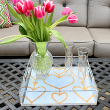 Load image into Gallery viewer, The Heartstring Acrylic Tray is square and has clear acrylic sides with two handles. It has a white and gold heart background on the bottom. It is styled with glasses and a vase of pink tulips. Heart art tray.
