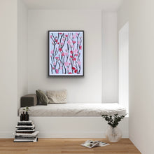 Load image into Gallery viewer, The Cherry Blossom Art Print is a giclee print printed on paper. It is shown displayed in a black frame over a day bed. This print has shades of pink and red blooms on a dark gray and black branches. The background is light blue.  It has a white border. 
