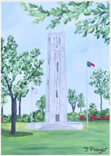 Load image into Gallery viewer, NC State Bell Tower Art on a Free Standing Acrylic Block styled on a coffee table beside a vase of orchids. This is a vertical landscape artwork of NCSU Memorial Belltower reproduced on an acrylic block. The belltower is in grassy field with blooming trees and shubbery. It is a small 5 x 7 inch block perfect to give for NC State graduation gift or alumni gift.