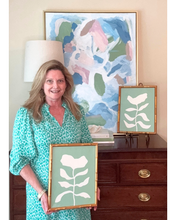 Load image into Gallery viewer, artist Jeanne Player holding a framed flower painting in a room with abstract paintings on the wall and a matching green and white flower art on the table.