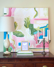 Load image into Gallery viewer, Decorative setup with a lamp, books, and a small gold framed lilac flower painting on a wooden surface in front of a colorful abstract painting with silver candlestick.