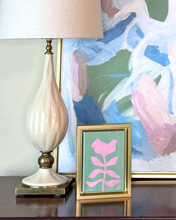 Load image into Gallery viewer, Decorative table setting with a lamp, framed modern pink flower on green background artwork, and small painting on a wood table.