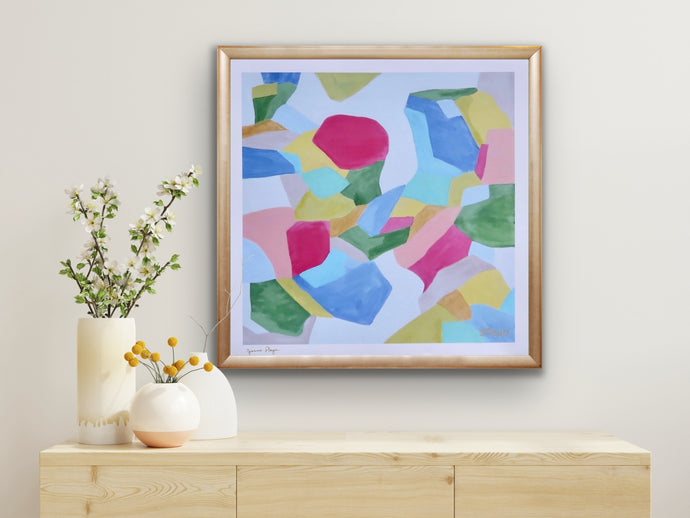 A colorful giclee art print on paper with a border. The artist has signed the front.  it is an abstract with shades of red, green, yellow, blue, aqua, peach, white and gray. It is shown in a gold frame over a natural wood table with vases of flowers.