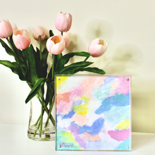 Load image into Gallery viewer, A colorful abstract painting on paper in a square acrylic frame. This painting has shades of blue, green, yellow, pink, fuchsia, orange and white. It is signed by the artist on the front.  It is displayed by a vase of flowers.
