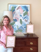 Load image into Gallery viewer, artist Jeanne Player holding a framed pink and white botanical artwork in front of a dresser with abstract art on the wall.
