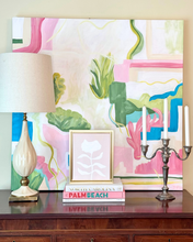 Load image into Gallery viewer, Palm Beach style interior with a lamp, books, and a framed pink and white floral artwork against a colorful abstract painting.
