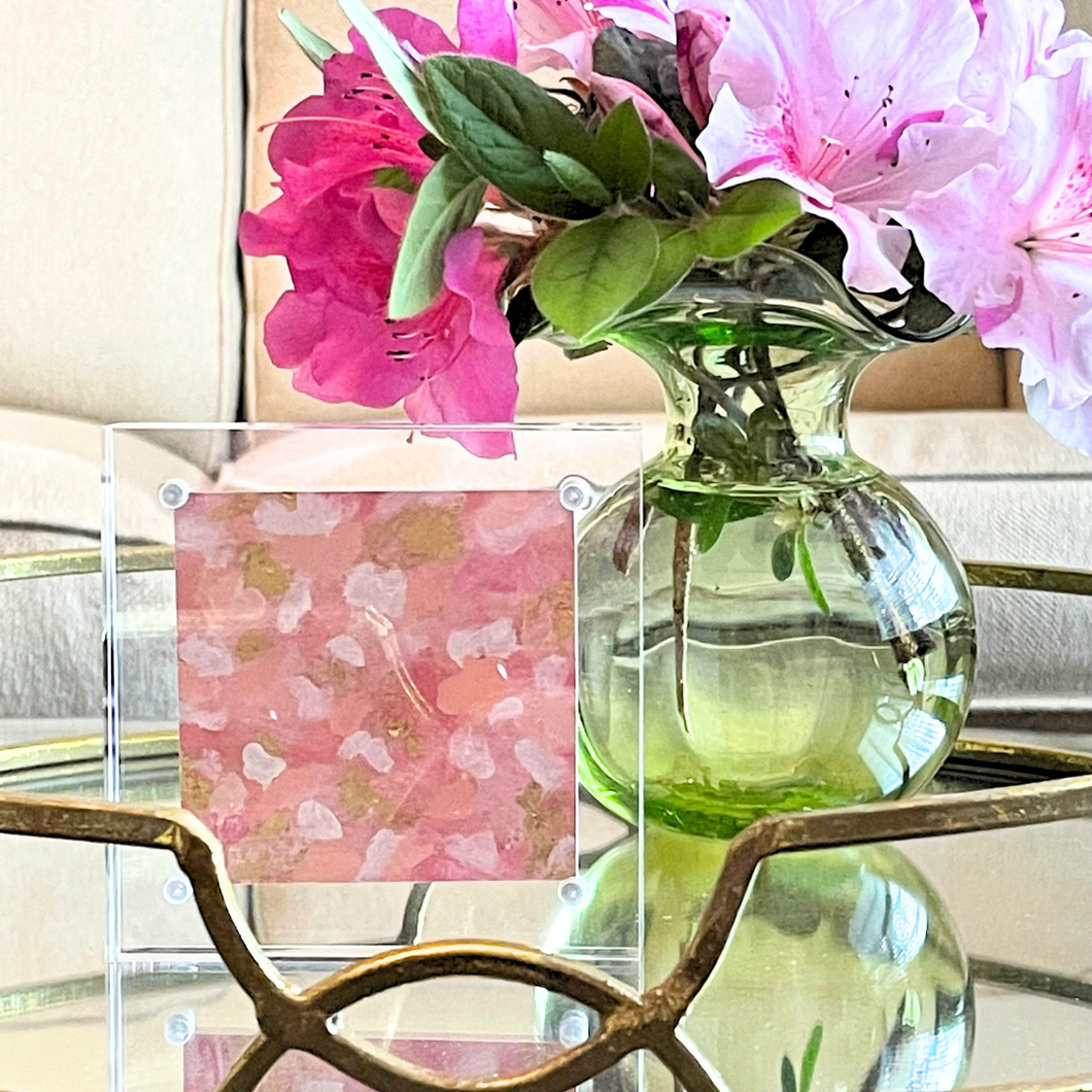 Small abstract painting in a clear acrylic block measuring 4 x 4 inches, The abstract looks like pink white and gold confetti. It is styled on a mirrored tray with a green vase holding pink azaleas.