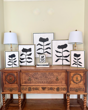 Load image into Gallery viewer, Wooden console table with decorative lamps and black floral paintings against a beige wall.
