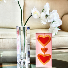 Load image into Gallery viewer, Heart Art. A 2. 5 x 7.5 vertical free standing acrylic block with a pink background and two red hearts outlined in gold connected by a gold line. It istyled on a mirrored tray beside a glass vase holding orchids.
