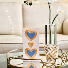 Load image into Gallery viewer, Two blue hearts outlined in gold with a gold heart between them. This original work of art is a shel sitter and has gold sides. The hearts are on a white background.
