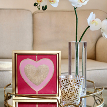Load image into Gallery viewer, Heart Art Acrylic on Canvas. Gold and pink hearts on red background. This square heart pop art painting comes in a gold frame and is dislplayed by a vase of orichids with a sofa in the background. Trending Maximalist art.
