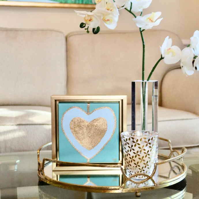 Heart Art on Canvas. Gold and blue hearts on a green background in a square gold frame. This is a modern style heart paintng. Colorful and happy hearts styled on a coffee table beside a vase of orchids  with a white sofa in the backround.