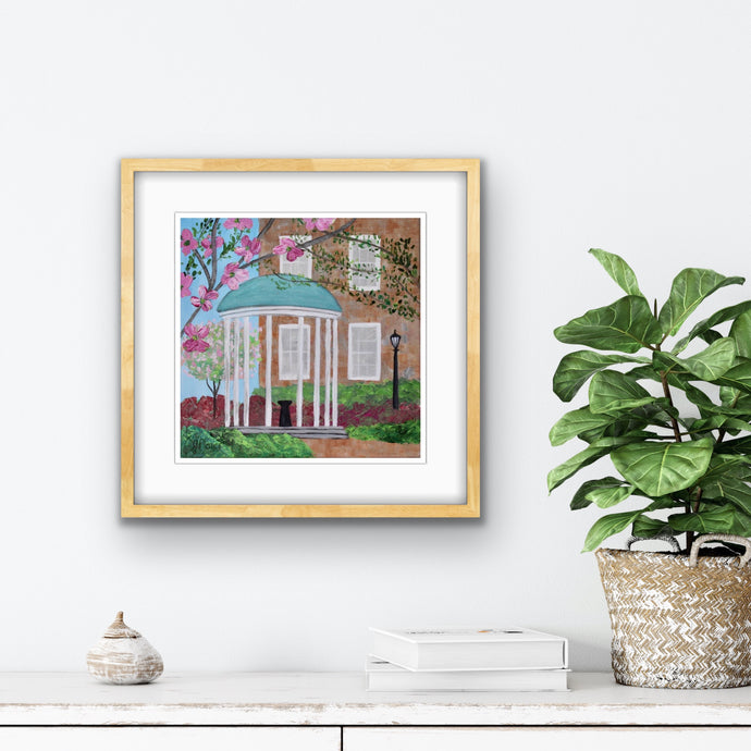 Colorful spring painting of the UNC Old Well. This landmark on the UNC campus is surrounded by blooming trees and flowers. There is a lamp post beside it with a brick building in the background. Flowering dogwoods, cherry blooms and azaleas. This is an abstract landscape print  shown framed above a white table with a green plant.