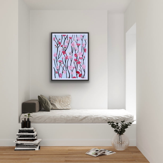 The Cherry Blossom Art Print is a giclee print printed on paper. It is shown displayed in a black frame over a day bed. This print has shades of pink and red blooms on a dark gray and black branches. The background is light blue.  It has a white border. 