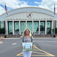 Load image into Gallery viewer, Artist Jeanne Player standing in front of Coleman Coliseum holding an original painting of the bulidling behind her.
