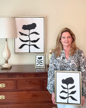 Load image into Gallery viewer, Artist Jeanne Player holding a framed black and white floral art piece in a room with a wooden chest, small black and white flower painting and a larger floral painting on the wall.and lamp.
