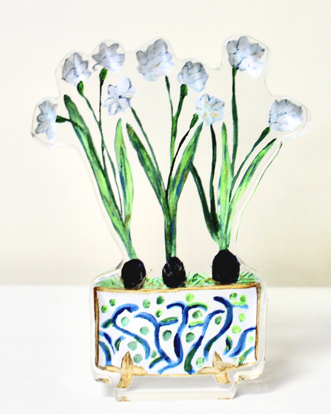 The paperwhite acrylic block is a colorful and fun work of art. This freestanding acrylic block art has three paperwhite bulbs with shades of white, yellow, green and brown. They are in a cute cachepot with a blue and green design gold edges and gold feet. This acrylic block art piece displays a vibrant arrangement of paperwhite flowers in a stylish cachepot, reproduced from original artwork by Jeanne Player. The freestanding design allows for versatile display, with the image visible from both sides.