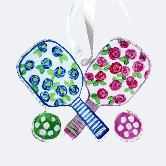 This pickleball ornament has a colorful floral design with shades of blue, green, pink and white. It is made of shatterproof acrylic and has a cut out shape. It is lightweight and has a white satin ribbon.