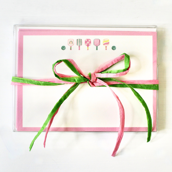 This is a cute and preppy stationery set of 10 flat note cards designed by Jeanne Player Fine Art. These cards have five colorful pickleball paddles and two green balls. The border is outlined in pink. The set has cute packaging with a pink and green raffia bow.