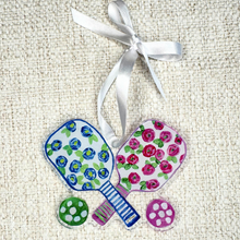 Load image into Gallery viewer, Pickleball Christmas ornament shaped like a paddle with whimsical floral artwork in pink, blue, rose, green, and white by Jeanne Player – lightweight acrylic holiday décor.
