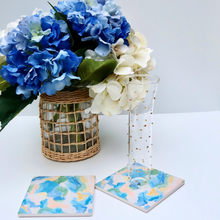 Load image into Gallery viewer, Square Abstract art coasters set of 2. These coasters have a cork back. They have shades of blue, green, white, yellow and coral. They are shown with a champagne glass and a vase of blue hydrangeas. 
