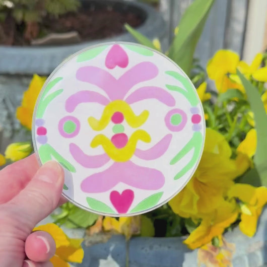 A video of a  colorful lucite coaster with an abstract art design. This video shows the front and back of the coaster and has yellow flowers in the background.