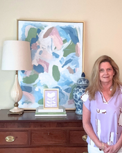 Load image into Gallery viewer, Artist Jeanne Player standing in a room with a decorative cabinet, lamp, and abstract painting.and a small lilac purple flower painting on canvas.
