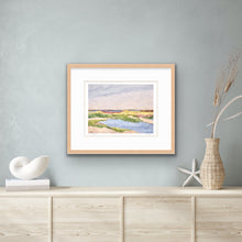 Load image into Gallery viewer, A colorful coastal marsh scene art print. This print has shades of tan, yellow, green, blue, brown and pink. It is signed on the front by the artist and shown framed  for display purposes over a table with a vase of flowers, and seashells.
