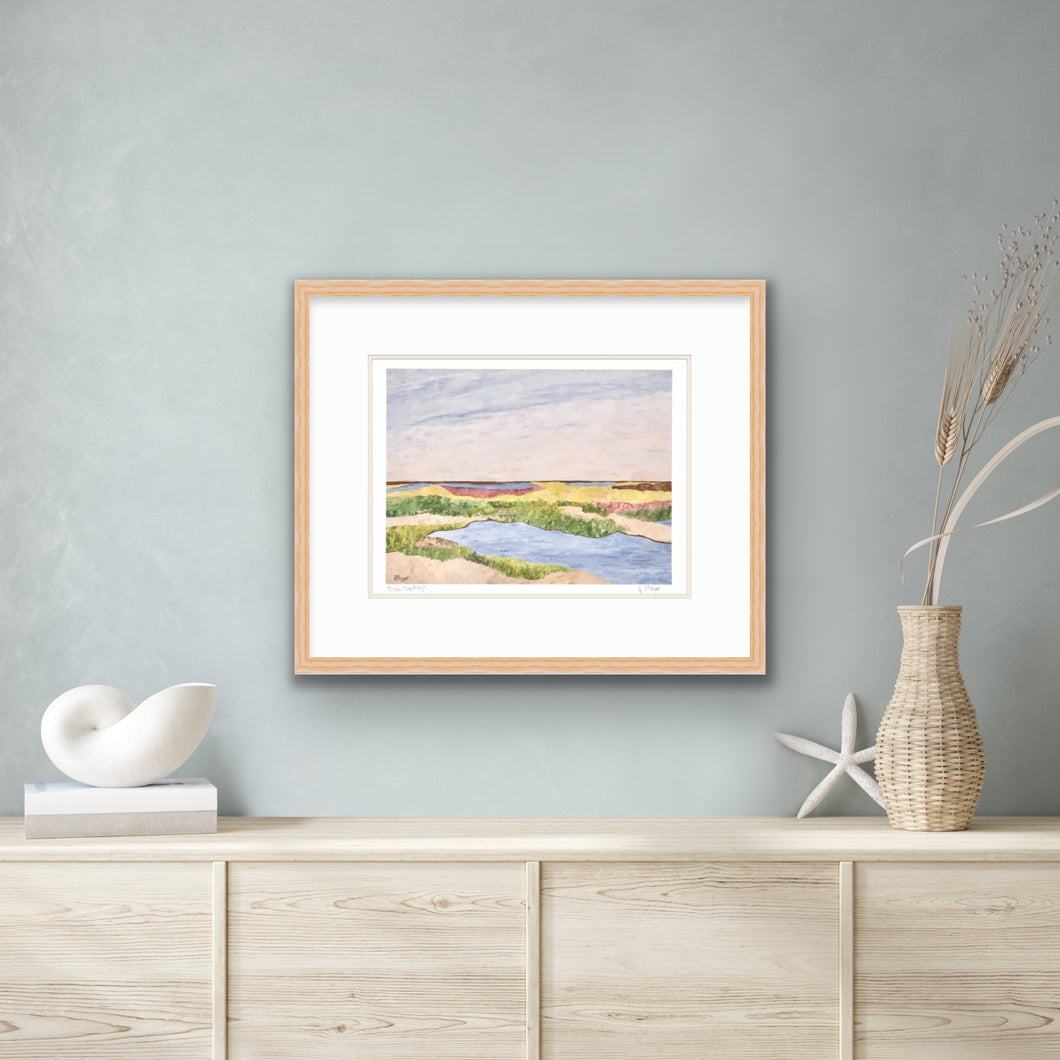 A colorful coastal marsh scene art print. This print has shades of tan, yellow, green, blue, brown and pink. It is signed on the front by the artist and shown framed  for display purposes over a table with a vase of flowers, and seashells.